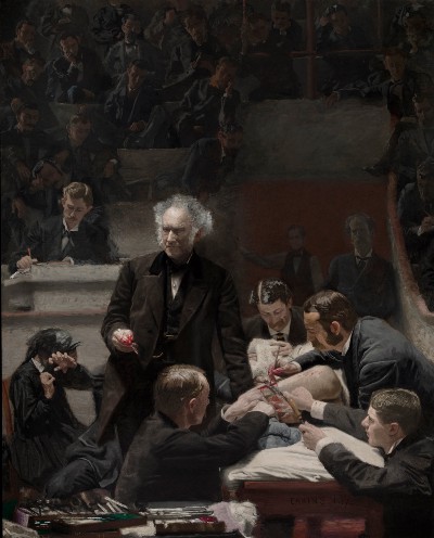 Surgery in a lecture theater, with the doctors wearing formal black suits. One doctor is turned to the audience, explaining what is happening.