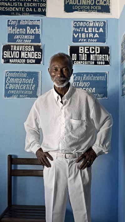 An older black gentleman wears a white button-down shirt and white trousers.
