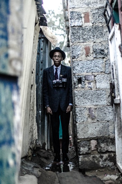A black man in a suit with a camera in an alley.