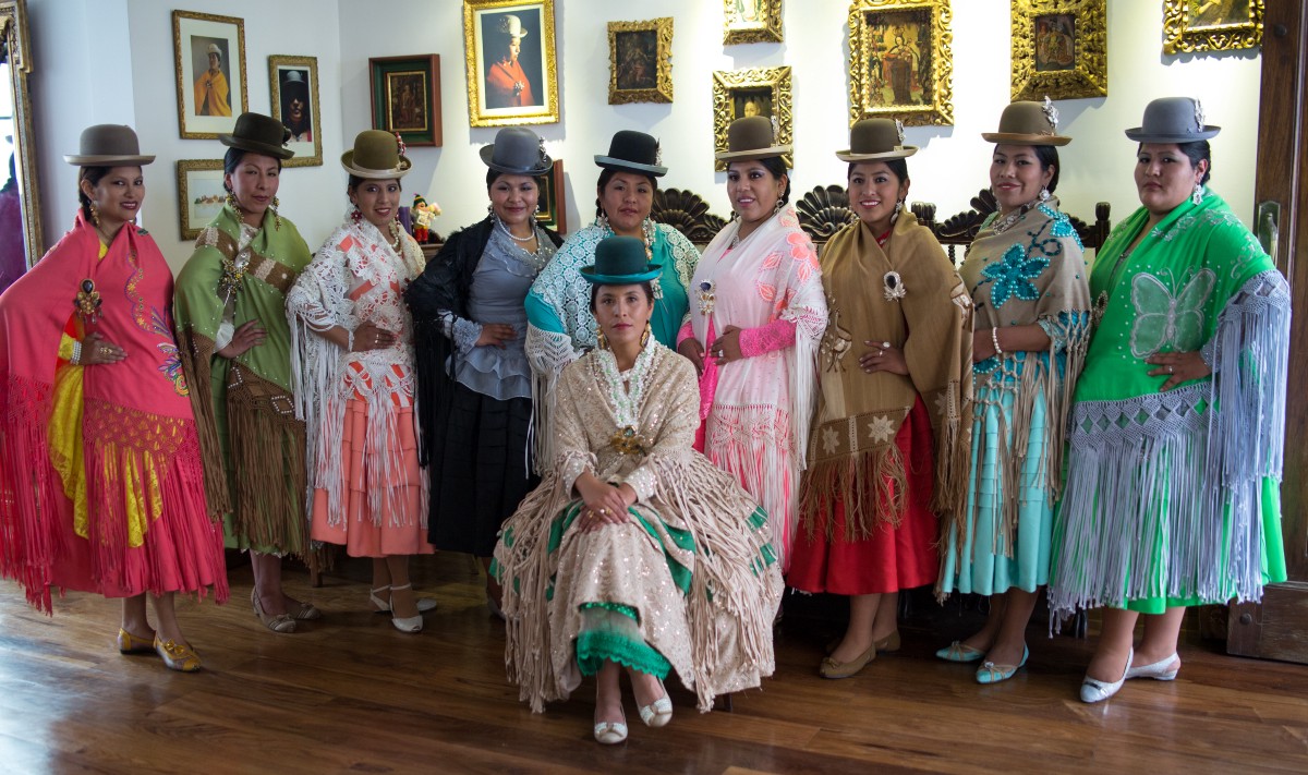 A group of Aymara women, all wearing traditional dress, pose for the camera in an art gallery.