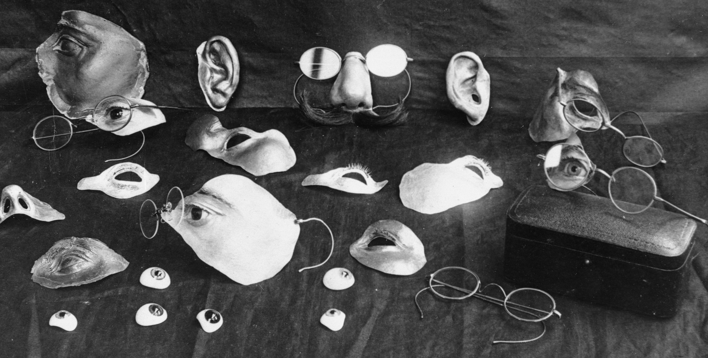 A black and white photo of a range of facial prosthetics–fake noses attached to glasses, fake cheeks, fake eyes, and fake ears.