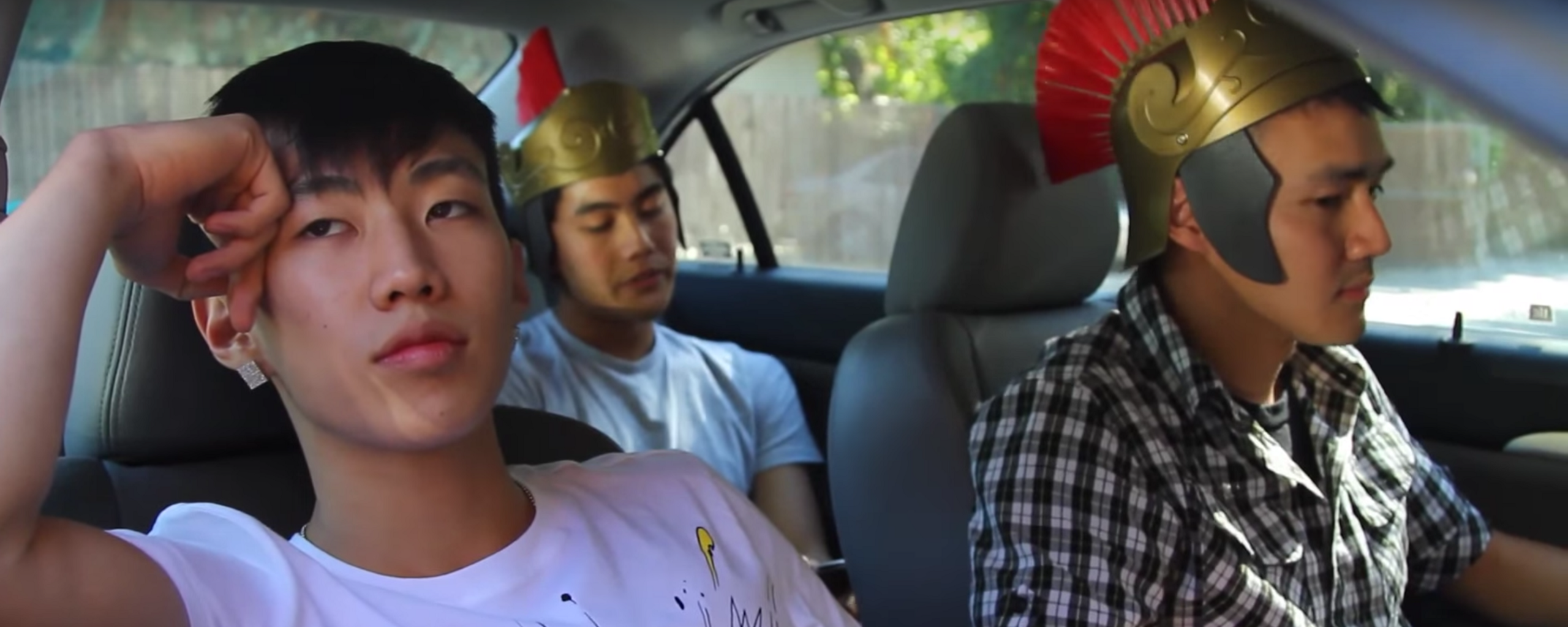 Three people sit in a car. Two wear plastic replicas of Roman legion helmets, while the other has his head on his hand.