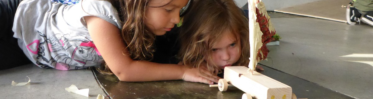 Two girls play with a wooden model car with a sail.