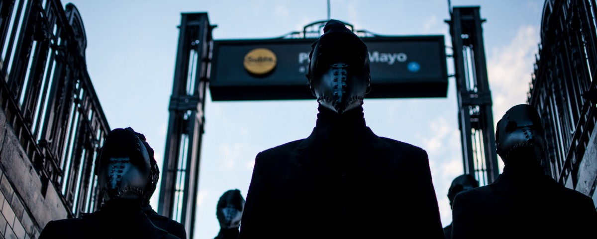 People stand still in formation in black suits, wearing black leather masks that completely hide their faces.