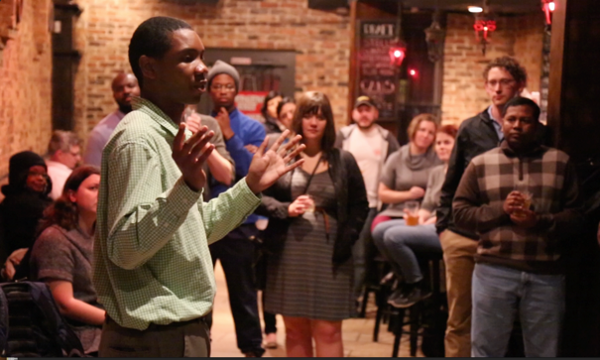 A young man gives a speech to a group of people in a crowded bar.