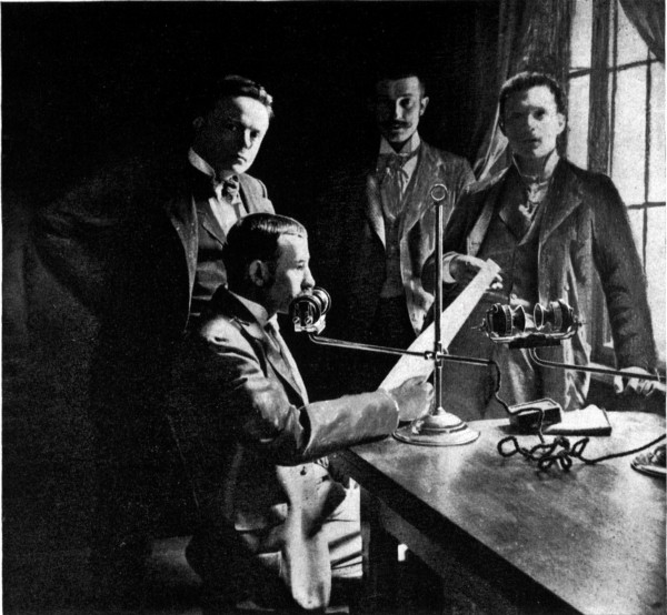 A group of men stand around a table, as one of their number reads into a primitive microphone from a piece of paper.