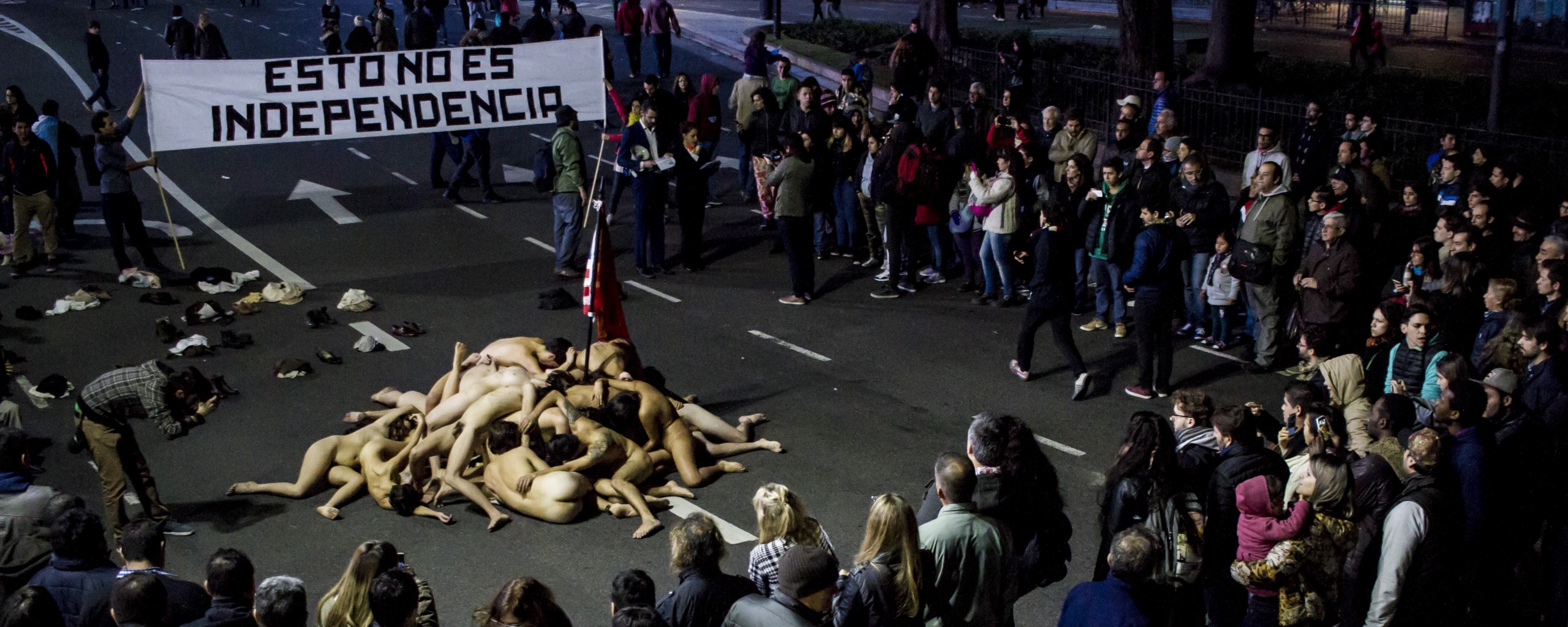A pile of naked protesters lie in a pile under a banner that reads ESTO NO ES INDEPENDENCIA.