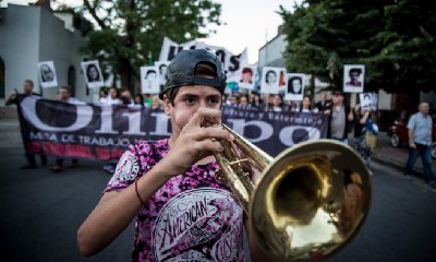 A protester marches in front of a banner, playing the trombone.