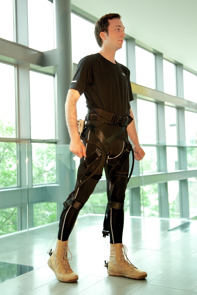 A man stands in a room, wearing an external supportive exosuit over his legs.
