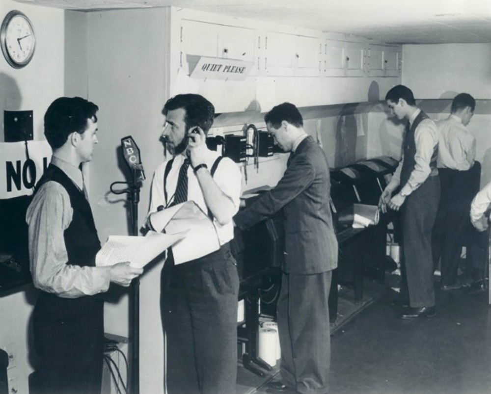 A row of white men stand next to radio broadcast equipment.