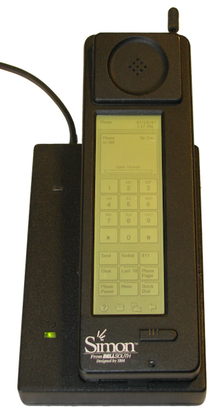 The IBM Simon, a large handset in a cradle. The front of the handset has a screen, displaying a range of apps as well as a keypad.