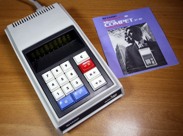A grey plastic calculator with instruction manual. The calculator has a power lead.