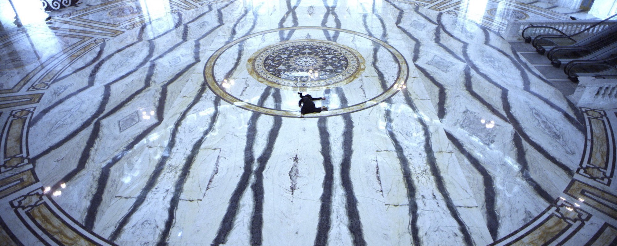 A figure in dark robes like sprawled in the center of a large, empty marble-floored gallery.