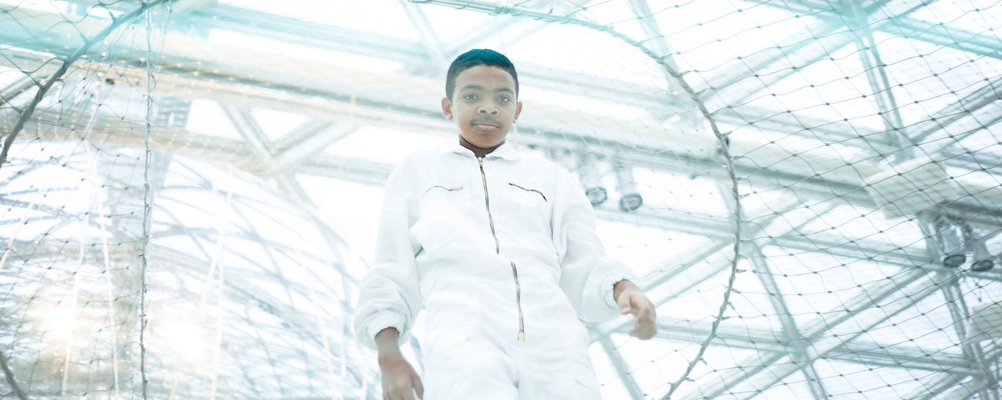 A young black boy in a white jumpsuit stands inside a large net-bubble structure.