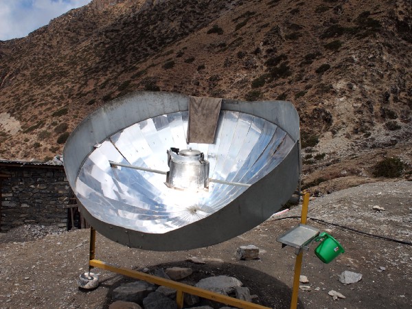 A large circular mirrored dish, standing on a brush-covered hillside, focuses sunlight onto a central chamber of water.