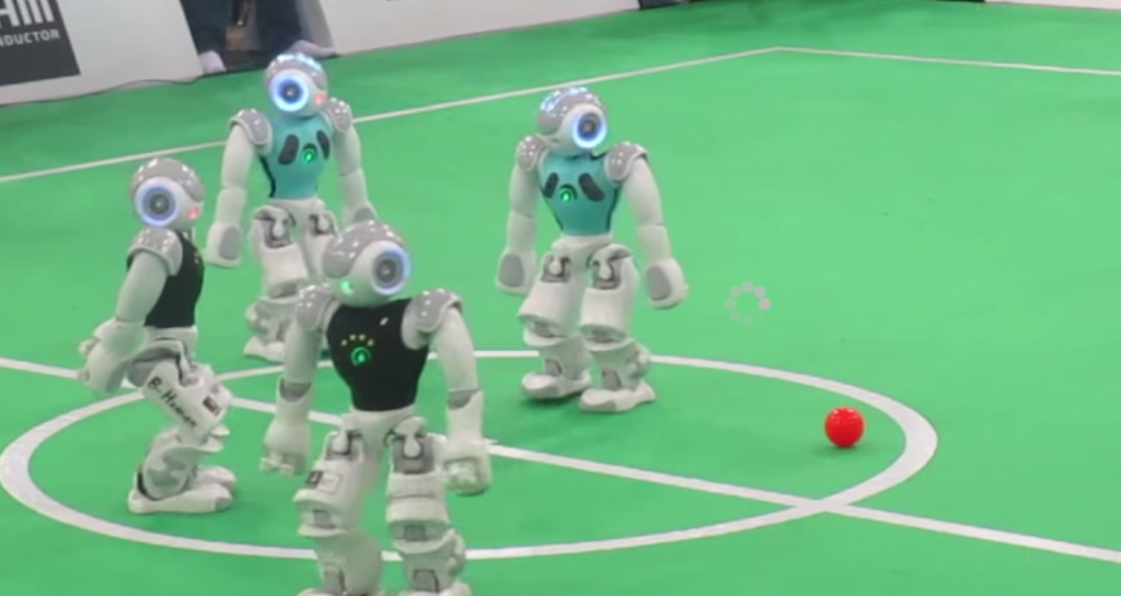 A group of small half-human height robots on a similarly scaled-down indoor soccer pitch, each moving towards a small orange ball.